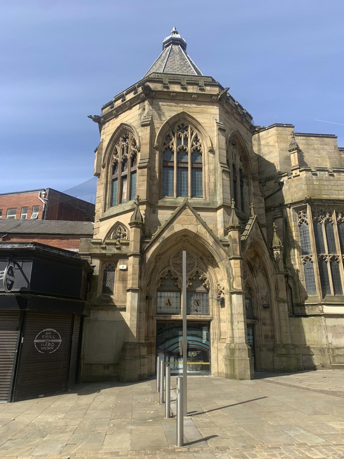 Photograph of a Victorian stone building. We see a tower like porch, very much looking like a cathedral porch.  It's hexagonal and we see the front door. To the left of the building is a modern brick built building. To the right is the subject building continuation.  It has ornate arch windows.  